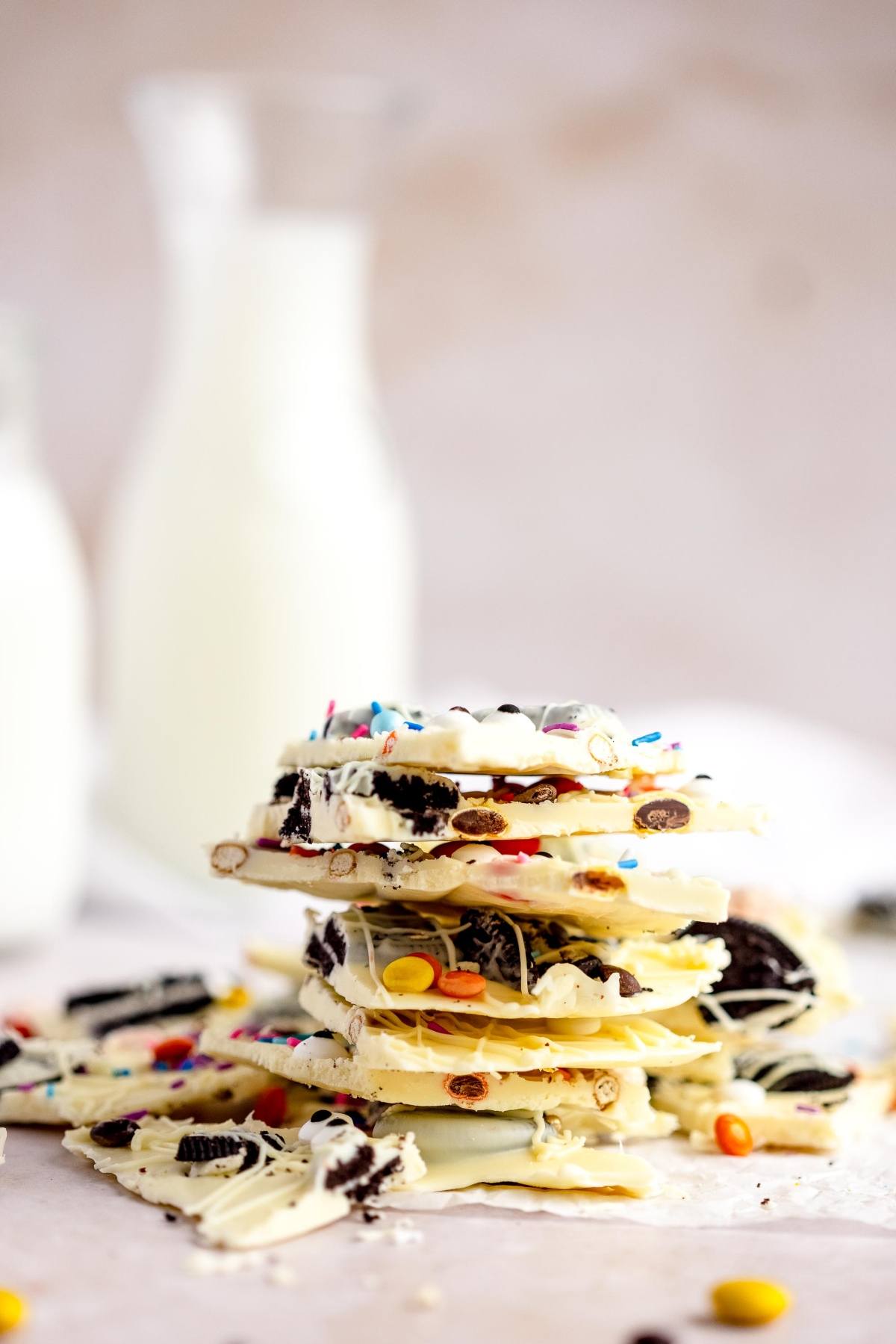 Stack of Halloween White Chocolate Candy Bark with cookies and colorful candies, with milk bottles blurred in the background—perfect for an easy candy bark recipe.