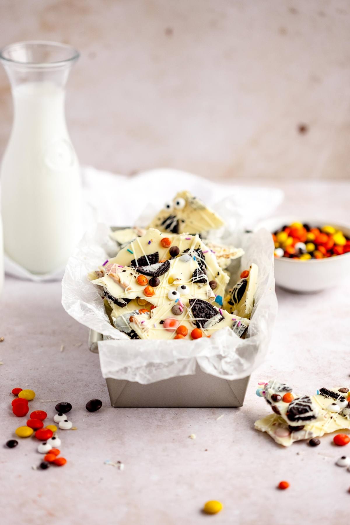 A tray of Halloween-themed white chocolate bark with candy and cookies, topped with googly monster eyes for a festive touch—an easy candy bark recipe perfect as a DIY Halloween treat, served beside milk and a bowl of candies.