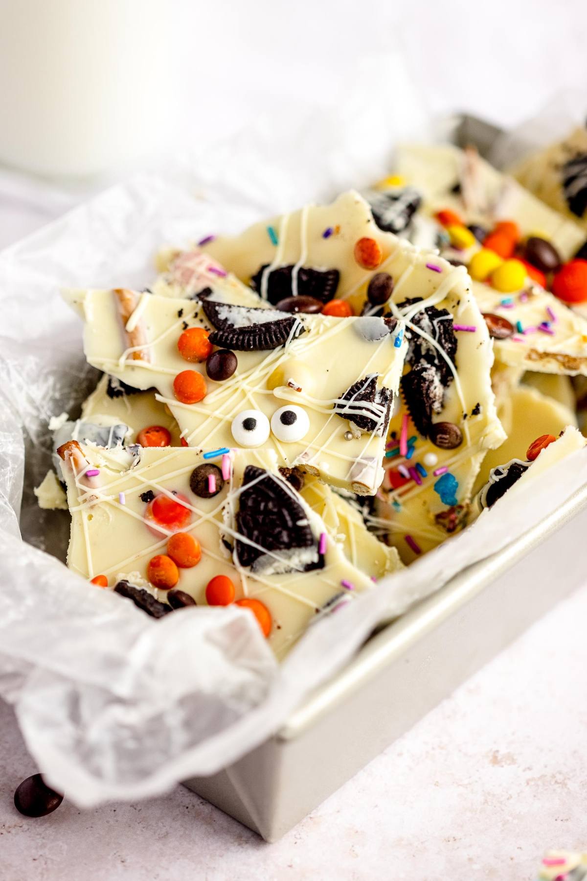 This Halloween White Chocolate Candy Bark features candy eyes, colorful sprinkles, and cookie pieces in a parchment-lined tin—an easy candy bark recipe perfect for spooky celebrations!.