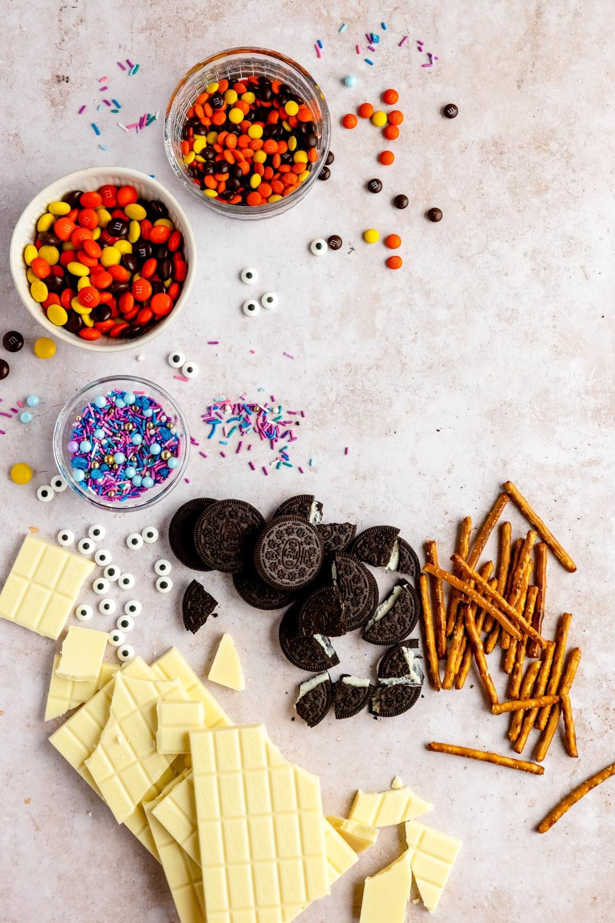 White chocolate, pretzel sticks, Oreos, candy eyes, sprinkles, and colorful candy in bowls on a beige surface—perfect ingredients for a DIY Halloween candy or a fun Halloween treat with googly monster eyes.