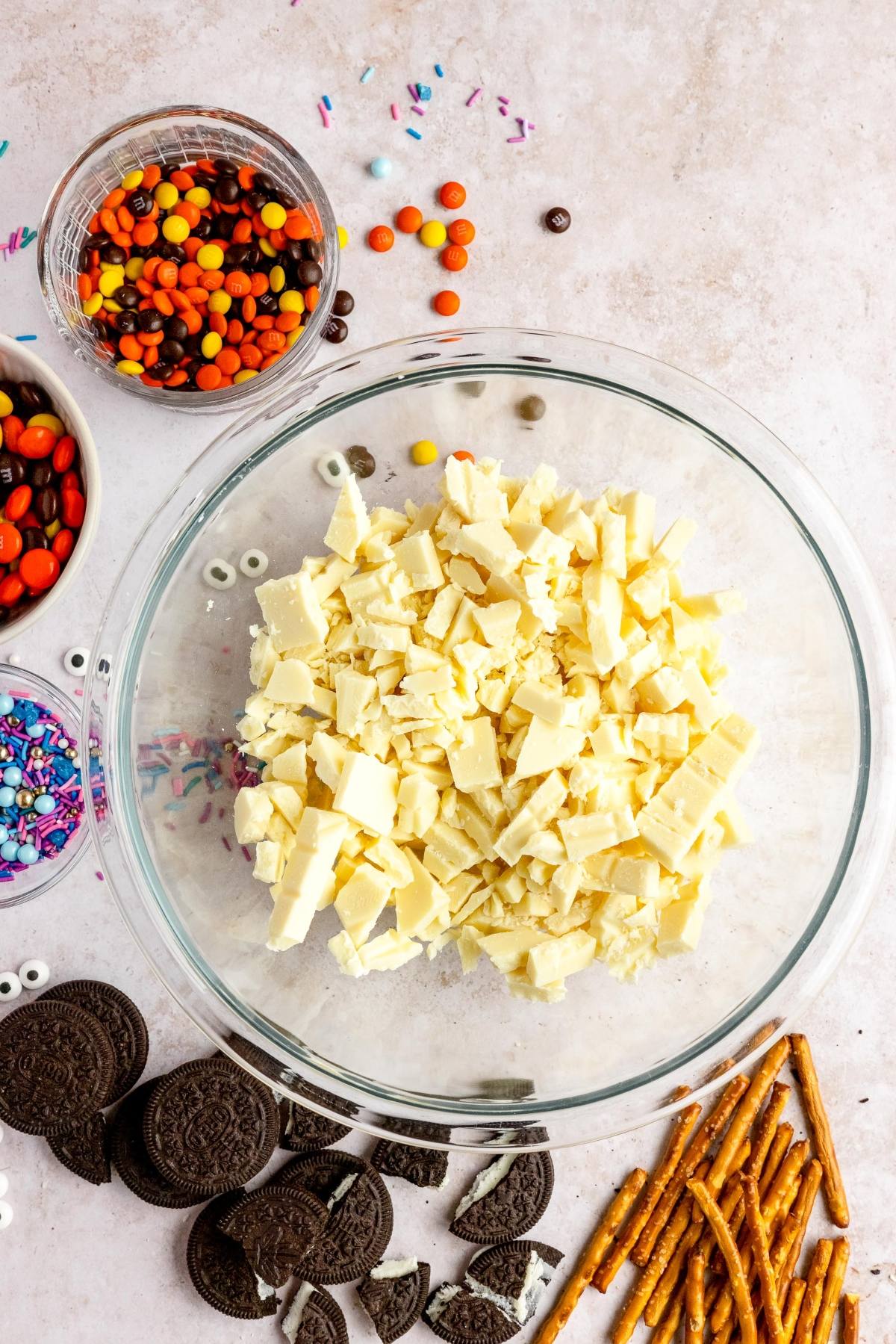 A bowl of chopped white chocolate surrounded by cookies, pretzels, candy, and colorful sprinkles—perfect for a fun DIY Halloween candy or as the base for an easy candy bark recipe.