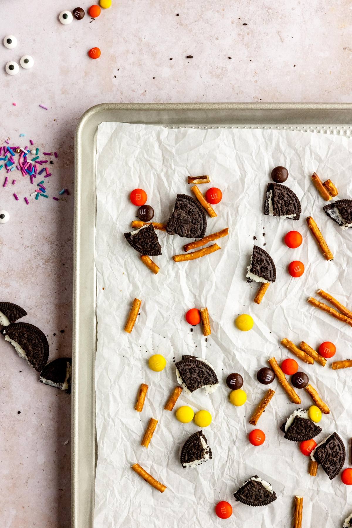 Baking sheet with broken Oreos, pretzel sticks, and colorful candy on parchment paper—an easy candy bark recipe perfect for Halloween treats with googly monster eyes, plus sprinkles nearby for extra fun.