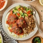 A plate with harissa meatballs, grain, salad, and sauce, surrounded by olives, dips, and more salad.