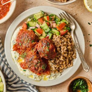 A plate with harissa meatballs, grain, salad, and sauce, surrounded by olives, dips, and more salad.