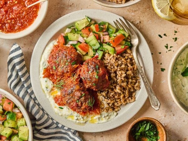 A plate with harissa meatballs, grain, salad, and sauce, surrounded by olives, dips, and more salad.