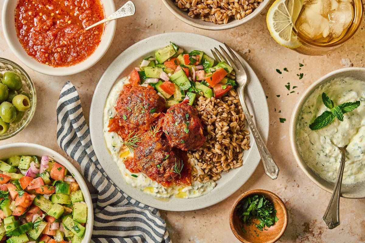 A plate with harissa meatballs, grains, cucumber salad, and sauces, surrounded by bowls of salad, olives, and drinks.