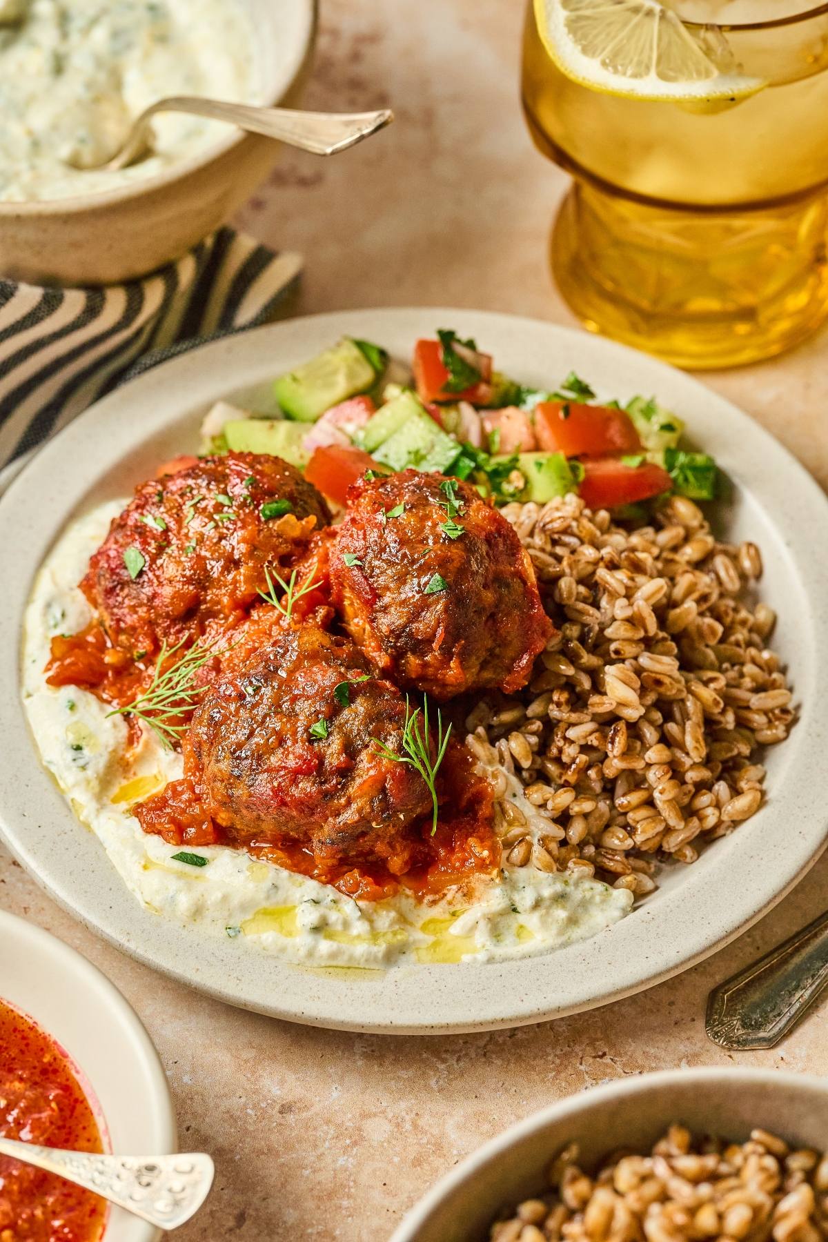 A plate with meatballs in tomato sauce, grains, salad, and tzatziki, next to a glass of iced tea with lemon.