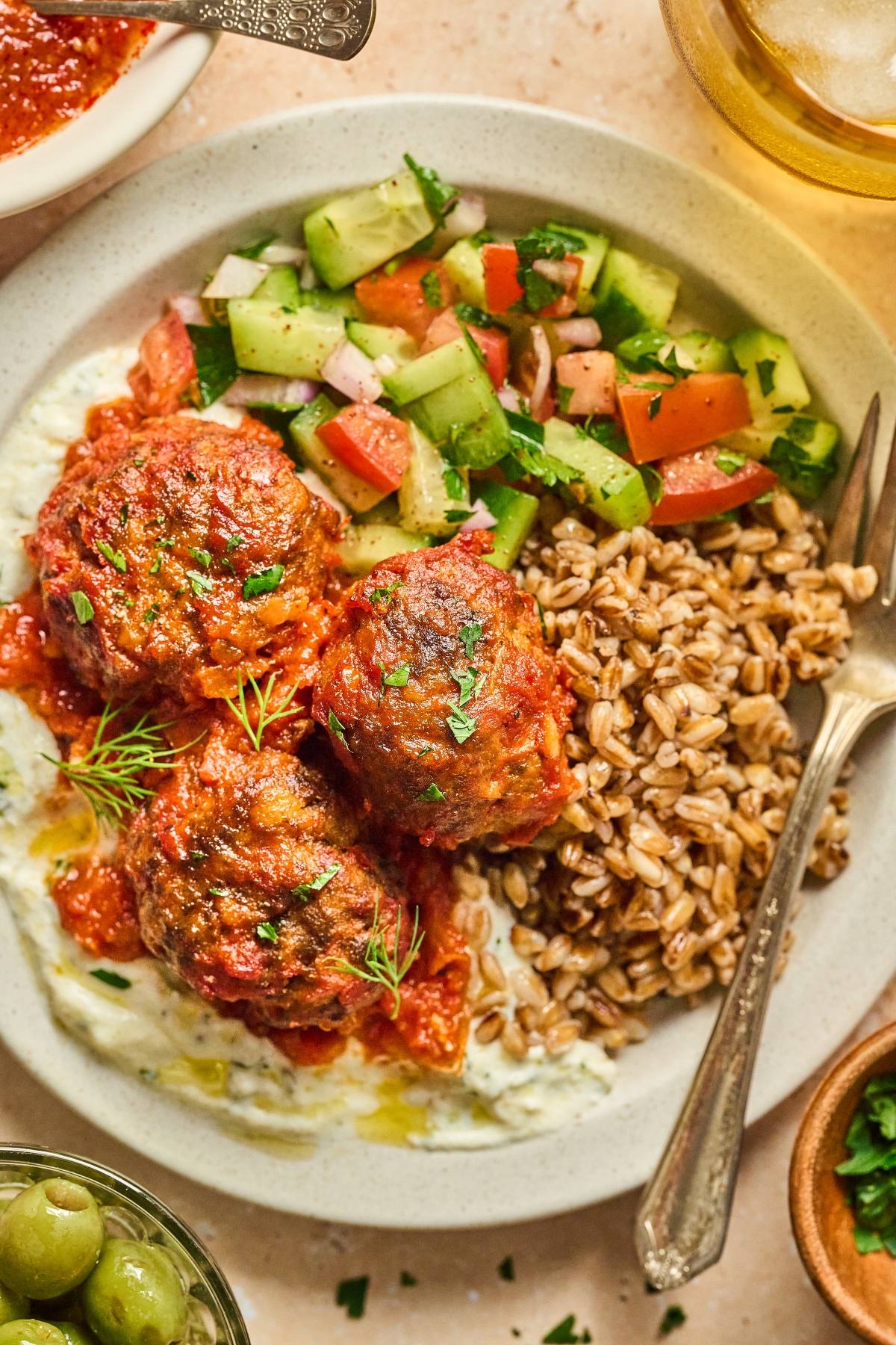 A plate with meatballs in tomato sauce, cucumber salad, cooked grains, and a creamy white sauce.