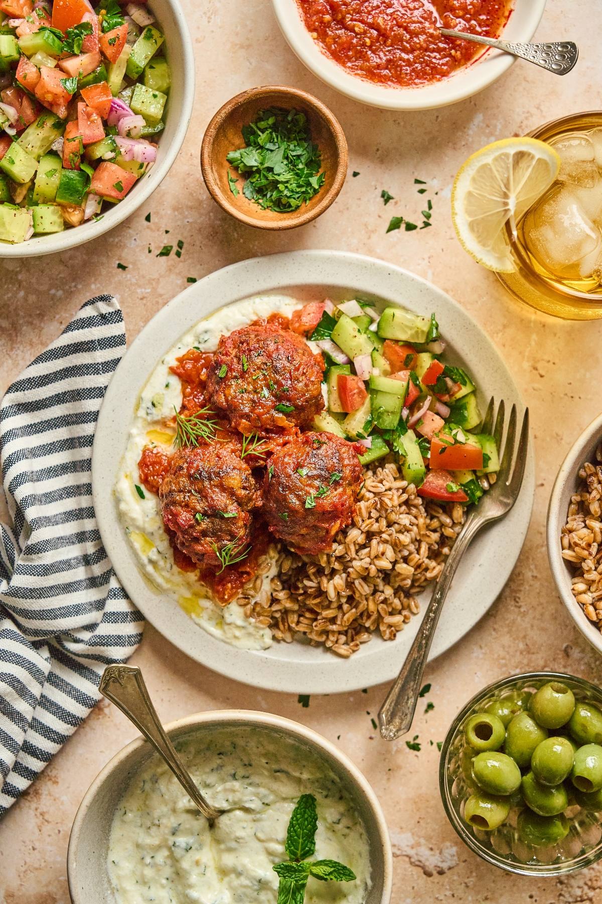 A plate with meatballs, rice, tzatziki, cucumber salad, and herbs, surrounded by bowls of sides and a drink.