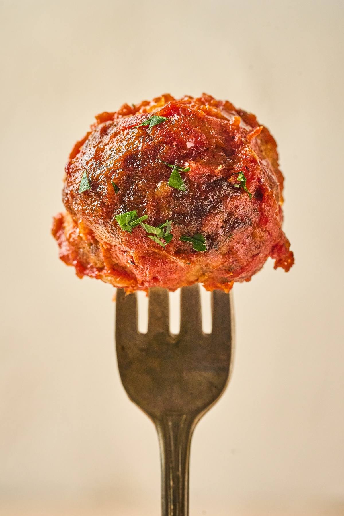 A single meatball with sauce and herbs on a fork, against a plain background.