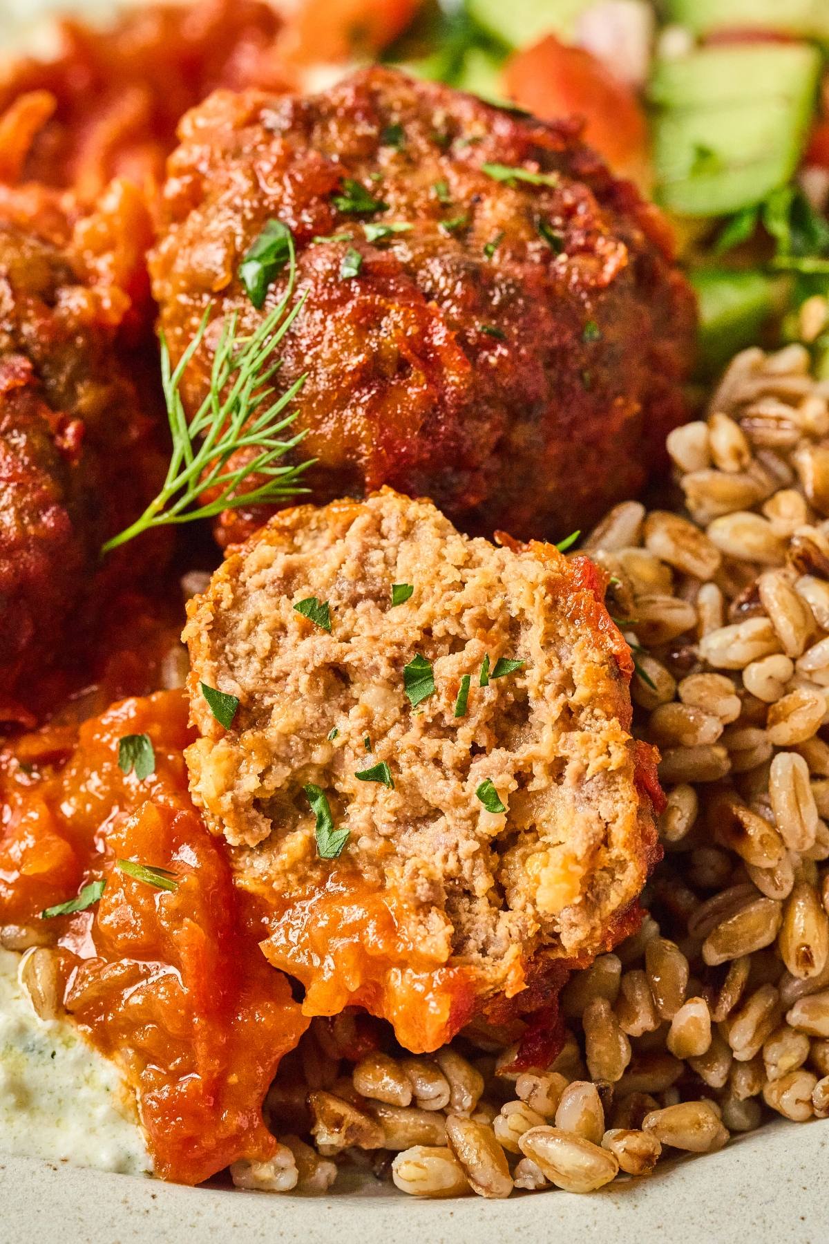 A close-up of a meatball with tomato sauce, cut open, served with grains and fresh vegetables.