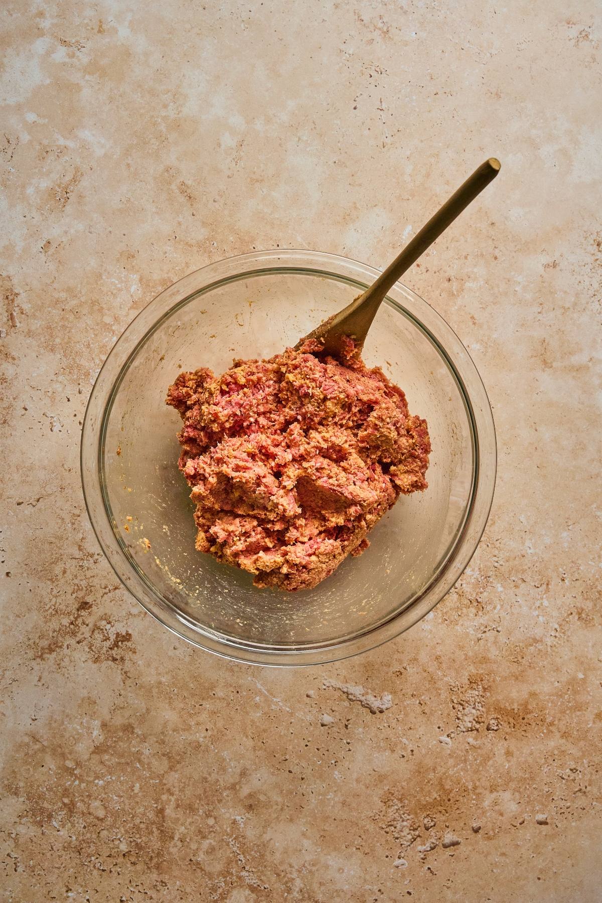 A glass bowl of ground meat mixture with a spoon on a light brown textured surface.