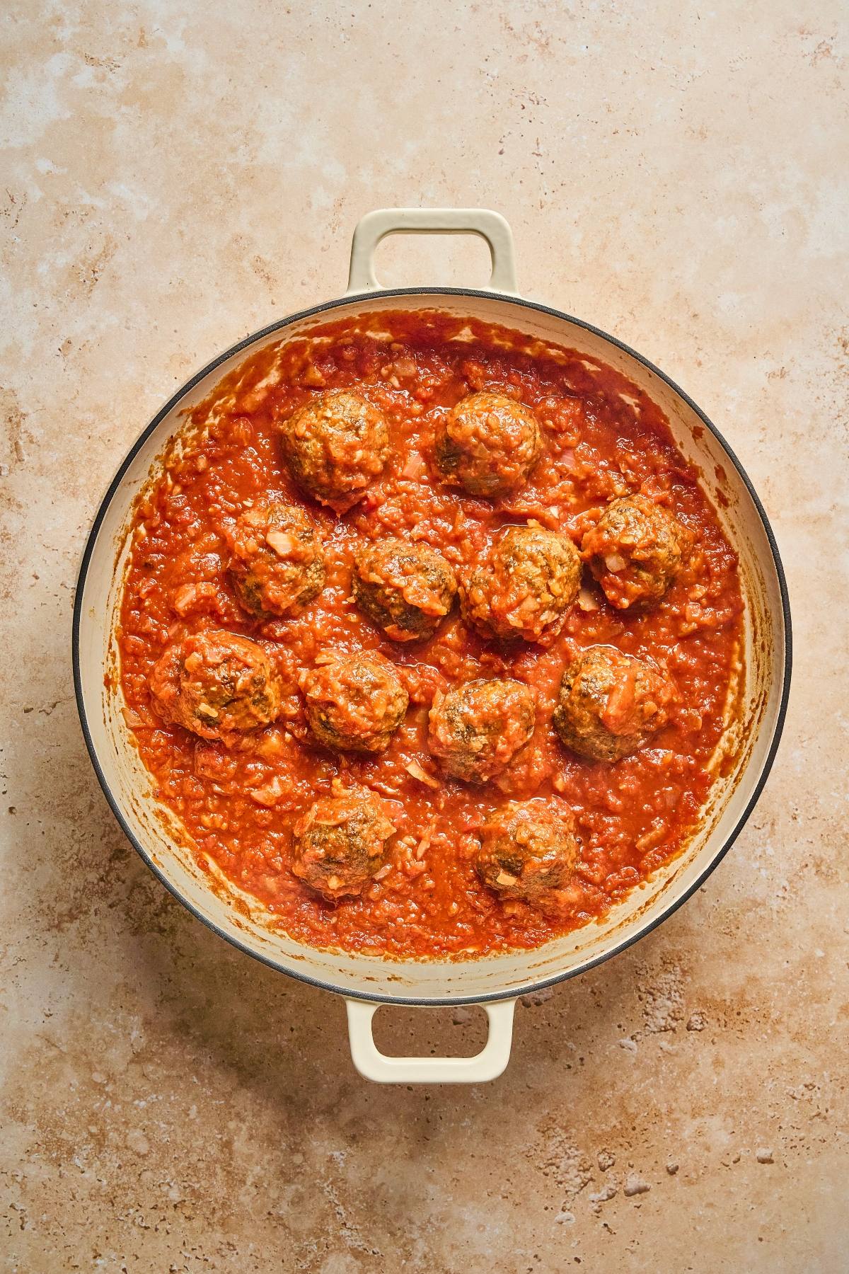Overhead view of a pot filled with meatballs simmering in tomato sauce on a light brown surface.