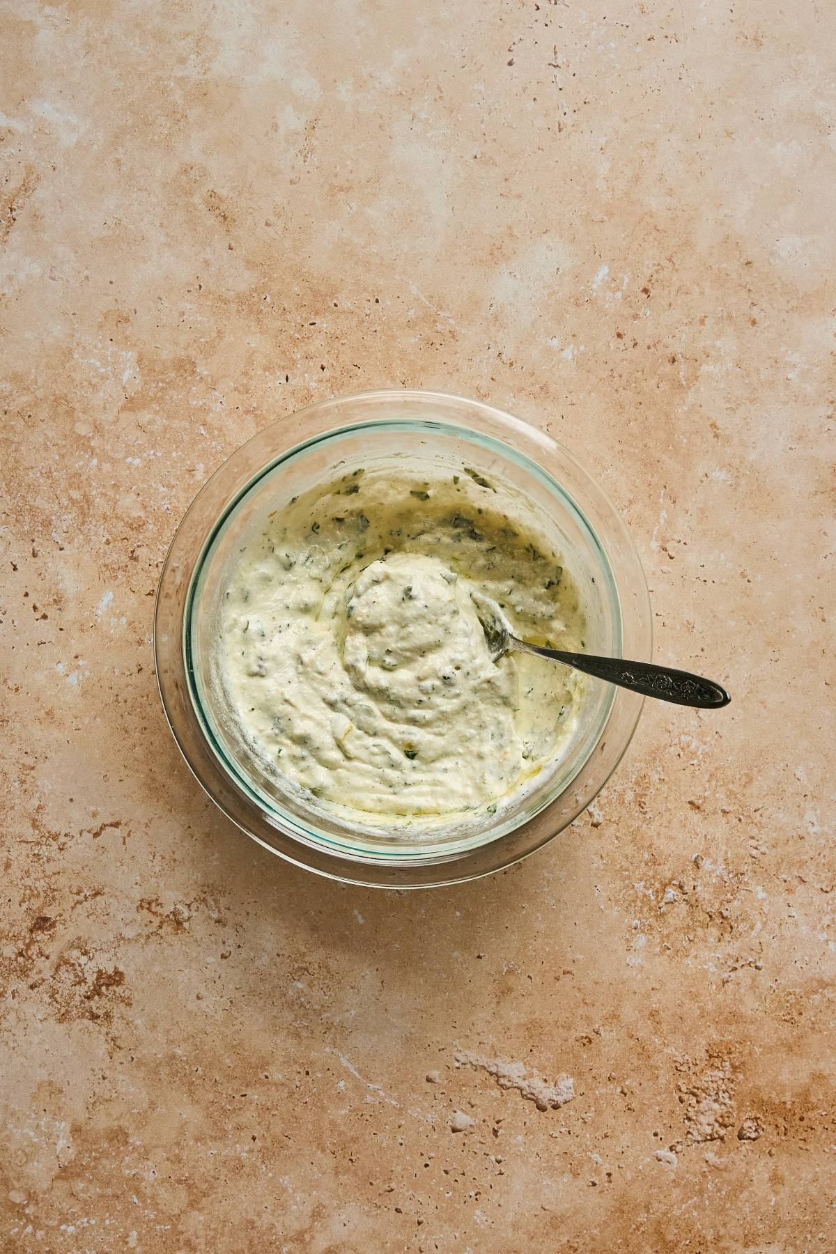 A glass bowl of creamy herb dip with a spoon, placed on a light brown textured surface.
