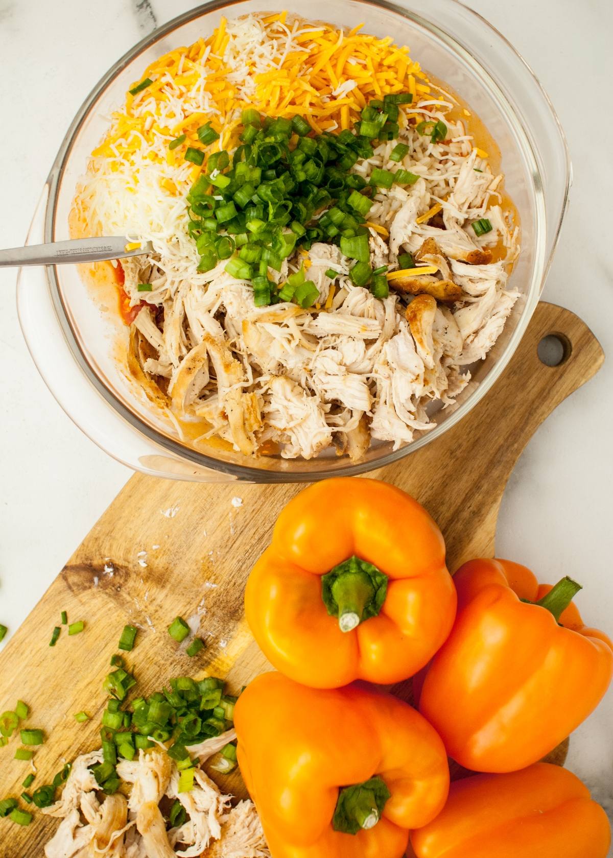 A glass bowl of shredded chicken, cheese, and green onions sits next to whole orange bell peppers on a cutting board—perfect for a fun Halloween dinner recipe.
