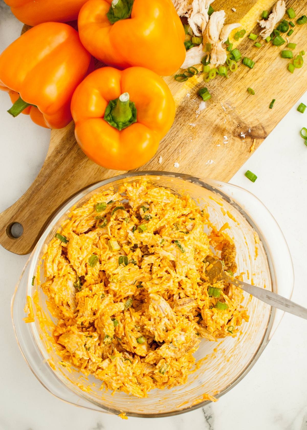 A glass bowl of shredded buffalo chicken dip sits beside orange jack o lantern peppers and sliced green onions—festive food to eat on Halloween arranged on a cutting board.