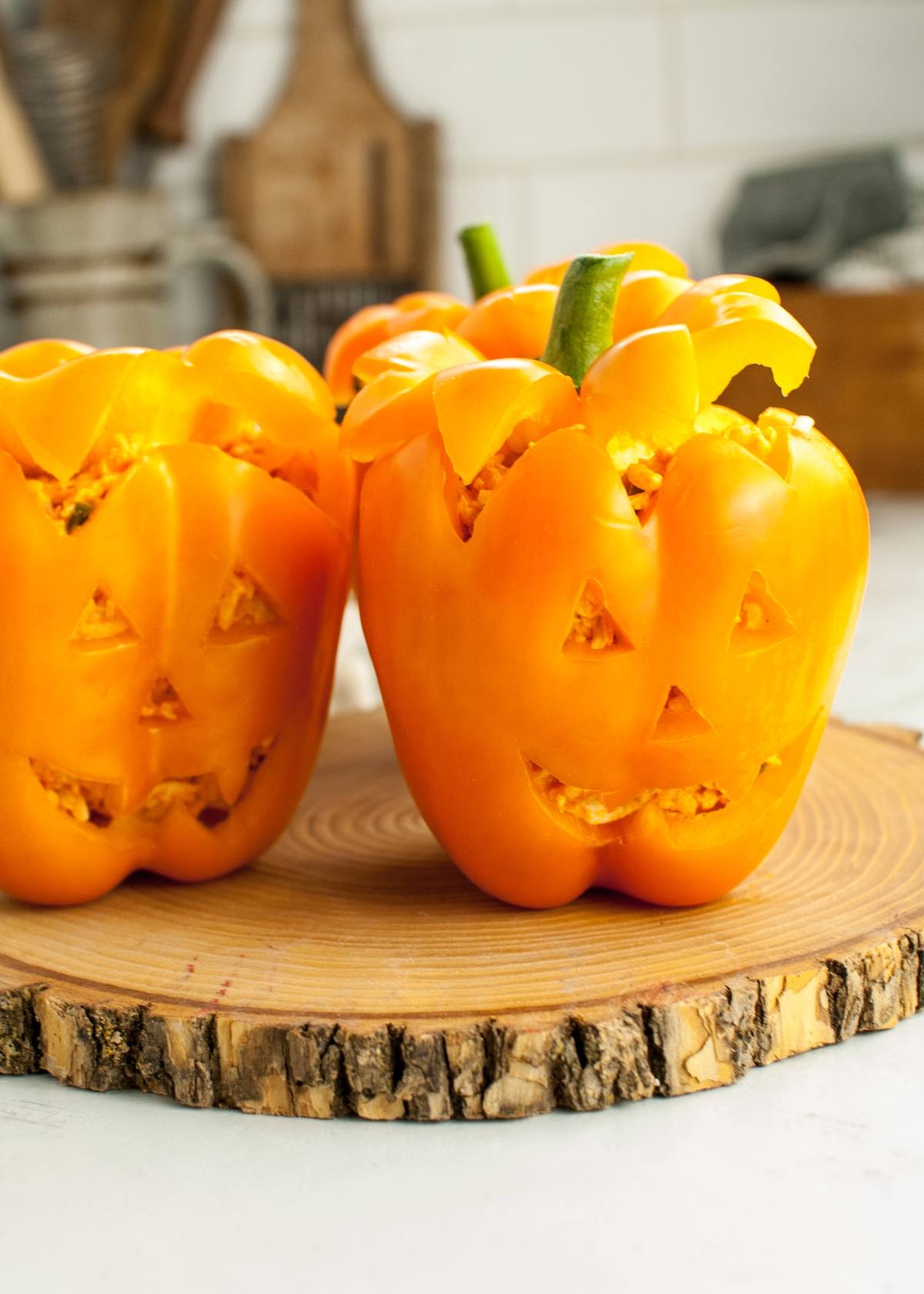 Two Jack-O-Lantern Stuffed Peppers, carved and smiling, sit on a wooden board—perfect food to eat on Halloween.
