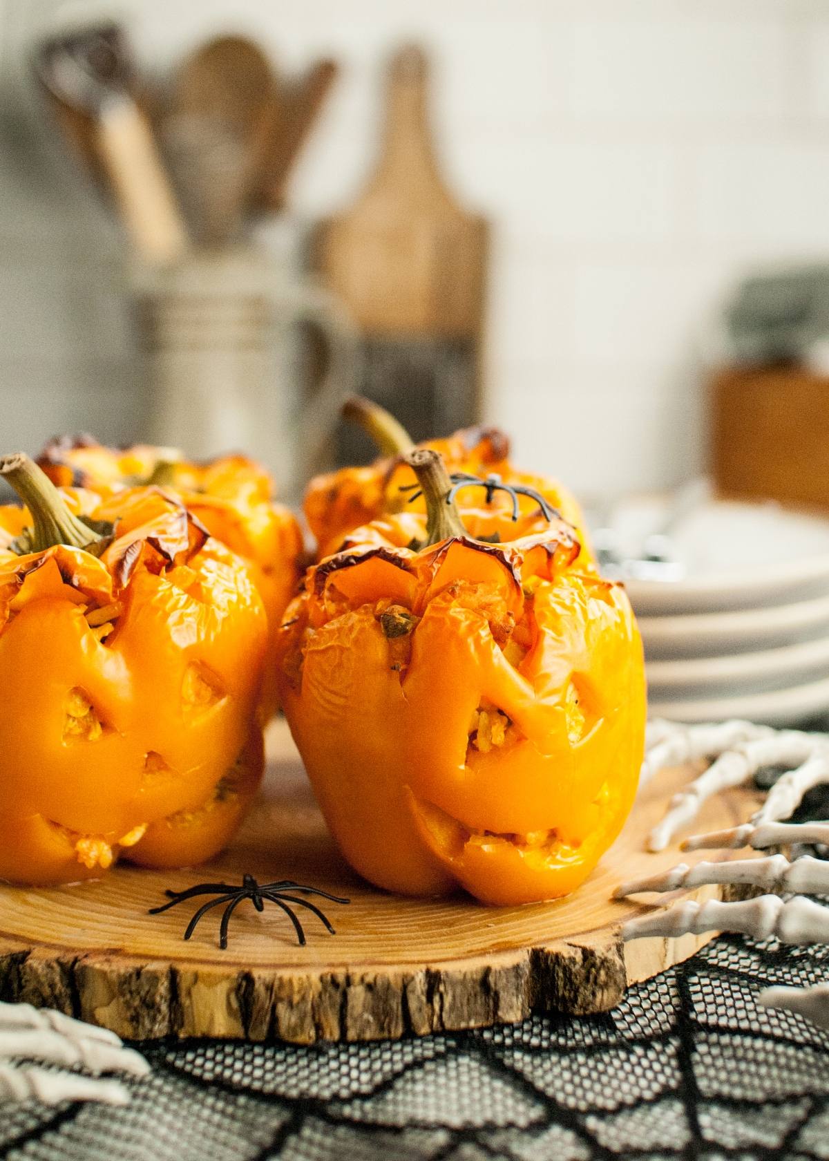 Three orange bell peppers carved like Jack-o'-lanterns, filled with a tasty Halloween dinner recipe, sit on a wooden board surrounded by festive Halloween decor—fun and delicious food to eat on Halloween.