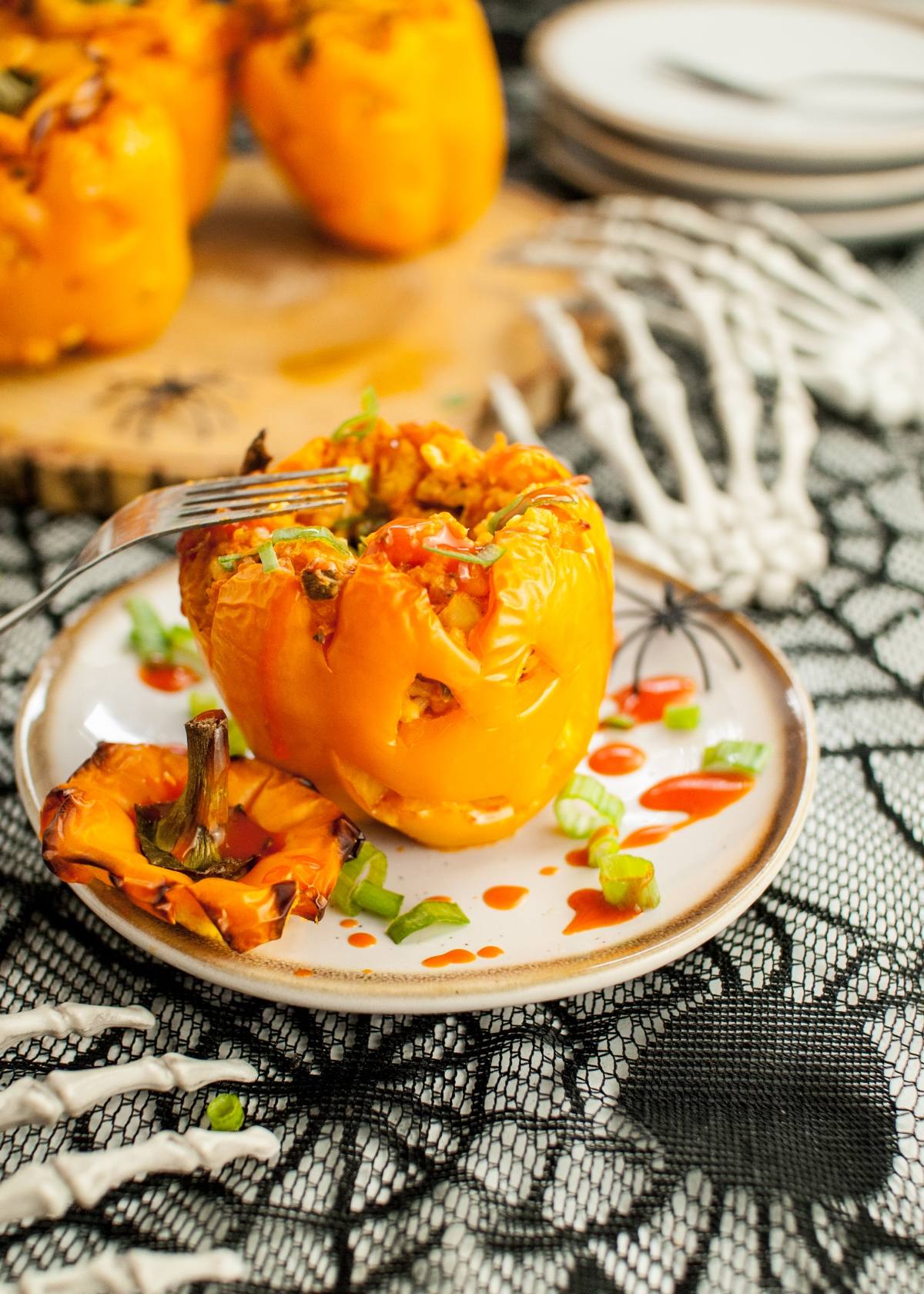 A Jack-O-Lantern Stuffed Pepper, topped with hot sauce, sits on a plate beside skeleton hands on a spiderweb tablecloth—an easy Halloween dinner recipe perfect for Halloween party ideas for adults.