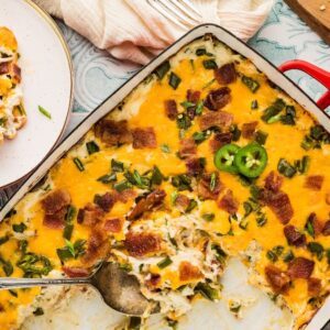Jalapeño popper casserole topped with bacon and extra jalapeños, served with a spoon on a decorative table.
