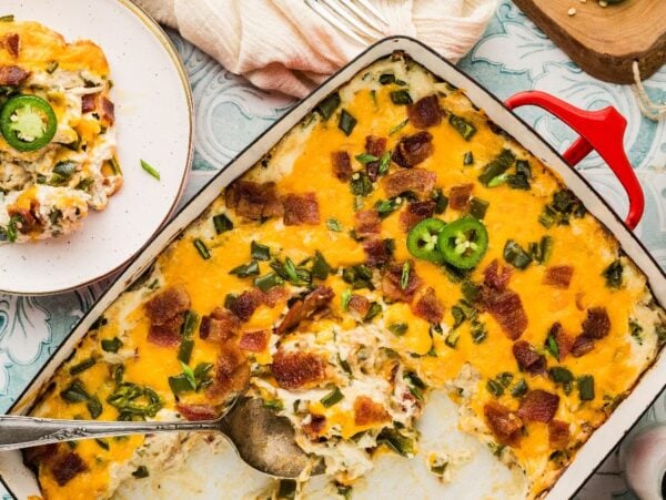 Jalapeño popper casserole topped with bacon and extra jalapeños, served with a spoon on a decorative table.