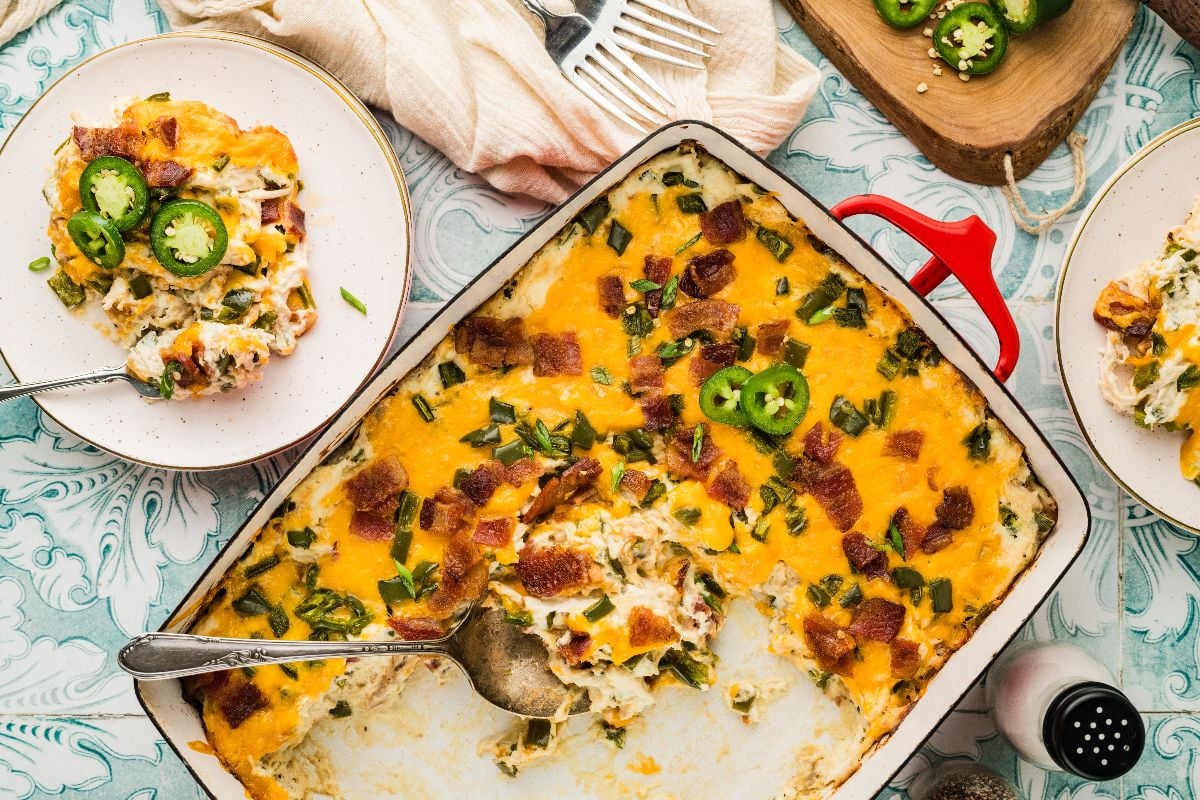 A cheesy jalapeño popper casserole baked in a red dish, served on a plate and topped with sliced jalapeños and green onions.