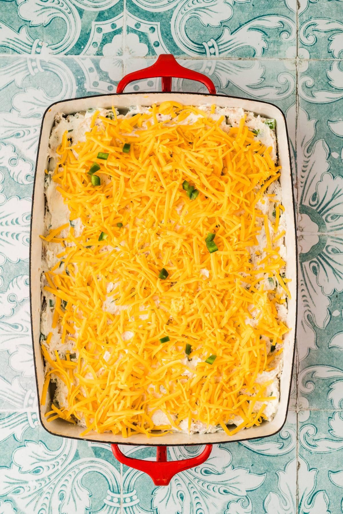 Rectangular baking dish with shredded cheddar cheese and diced green peppers on a creamy white layer.