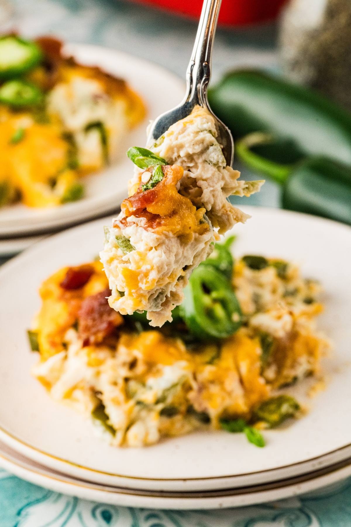A fork holding a cheesy, creamy casserole bite over a plate of the same dish, topped with bacon and jalapeños.