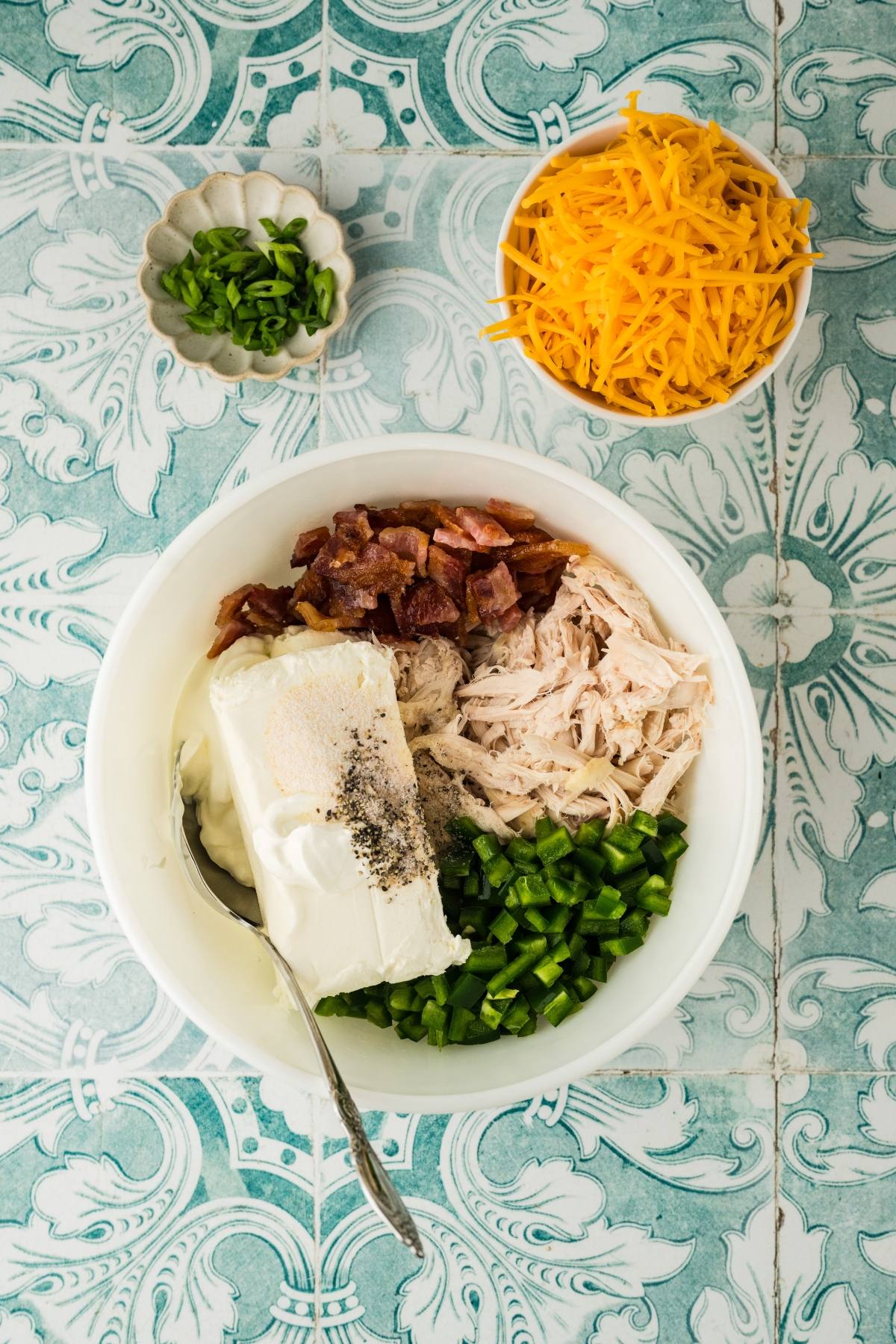 A bowl with cream cheese, shredded chicken, bacon, jalapeños, and seasonings, next to cheese and chopped onions.