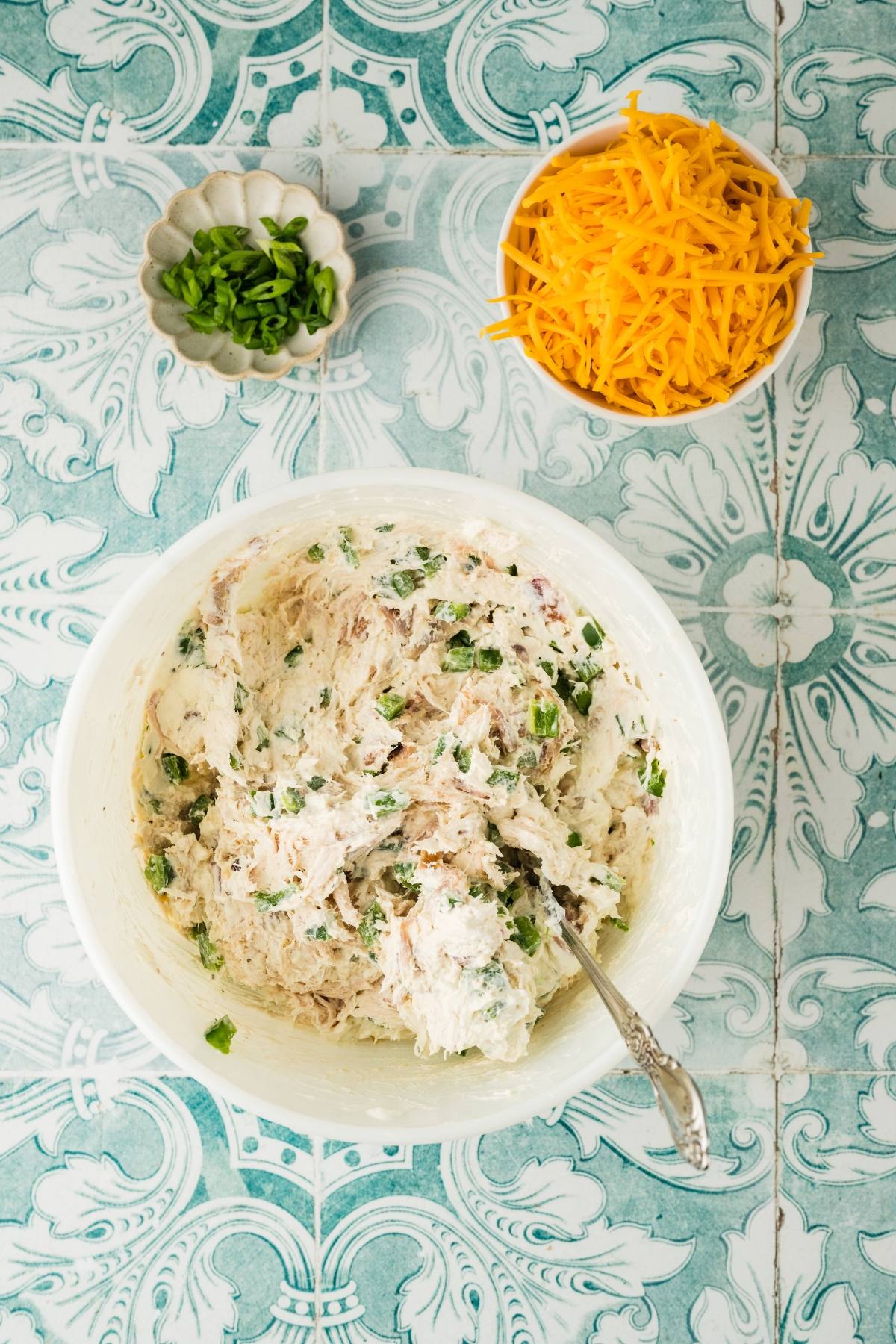 A bowl of chicken salad mix, with chopped green onions and shredded cheddar cheese on a patterned tile surface.