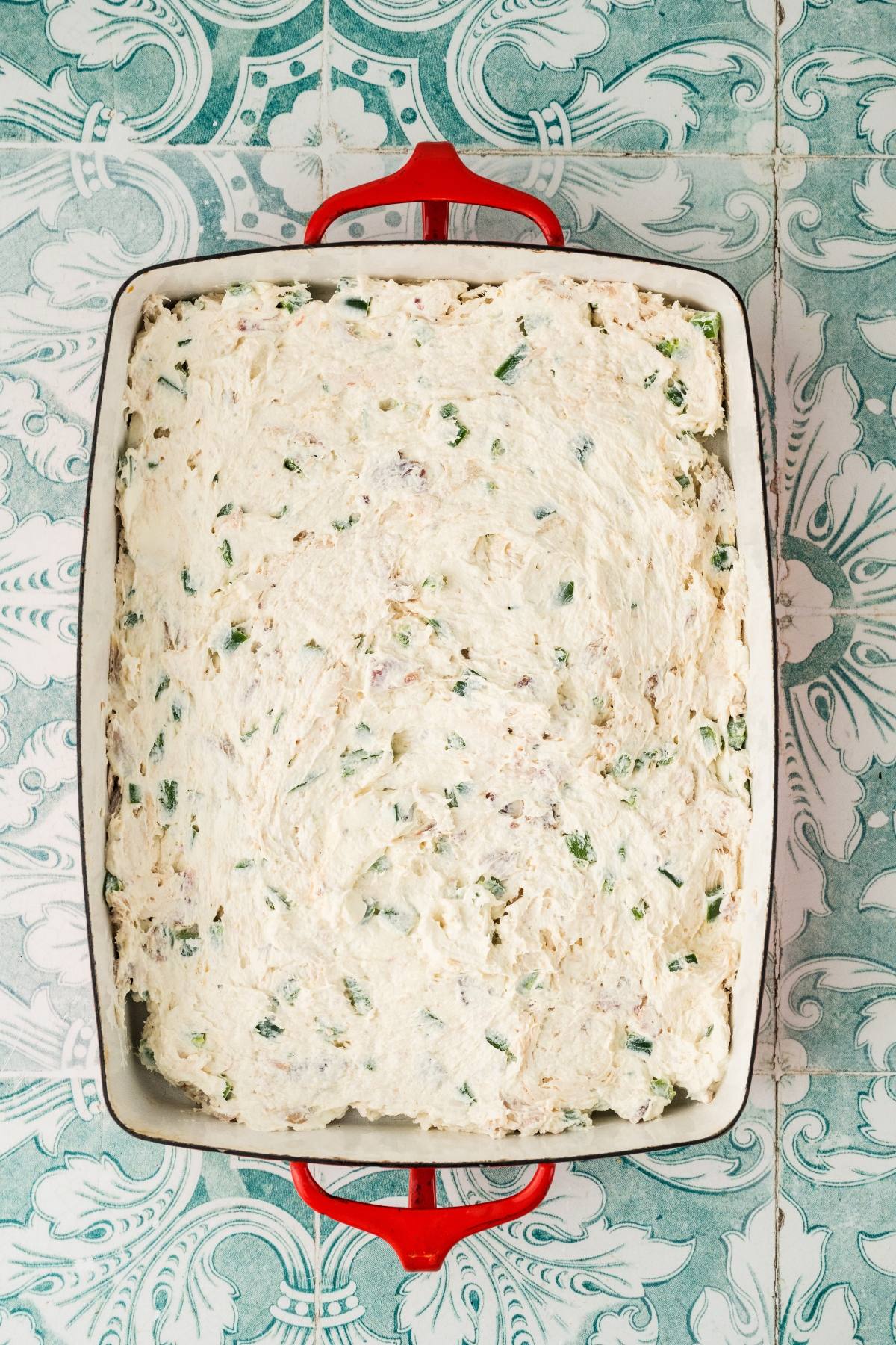 Rectangular baking dish with an unbaked creamy casserole topped with chopped green herbs on a patterned surface.