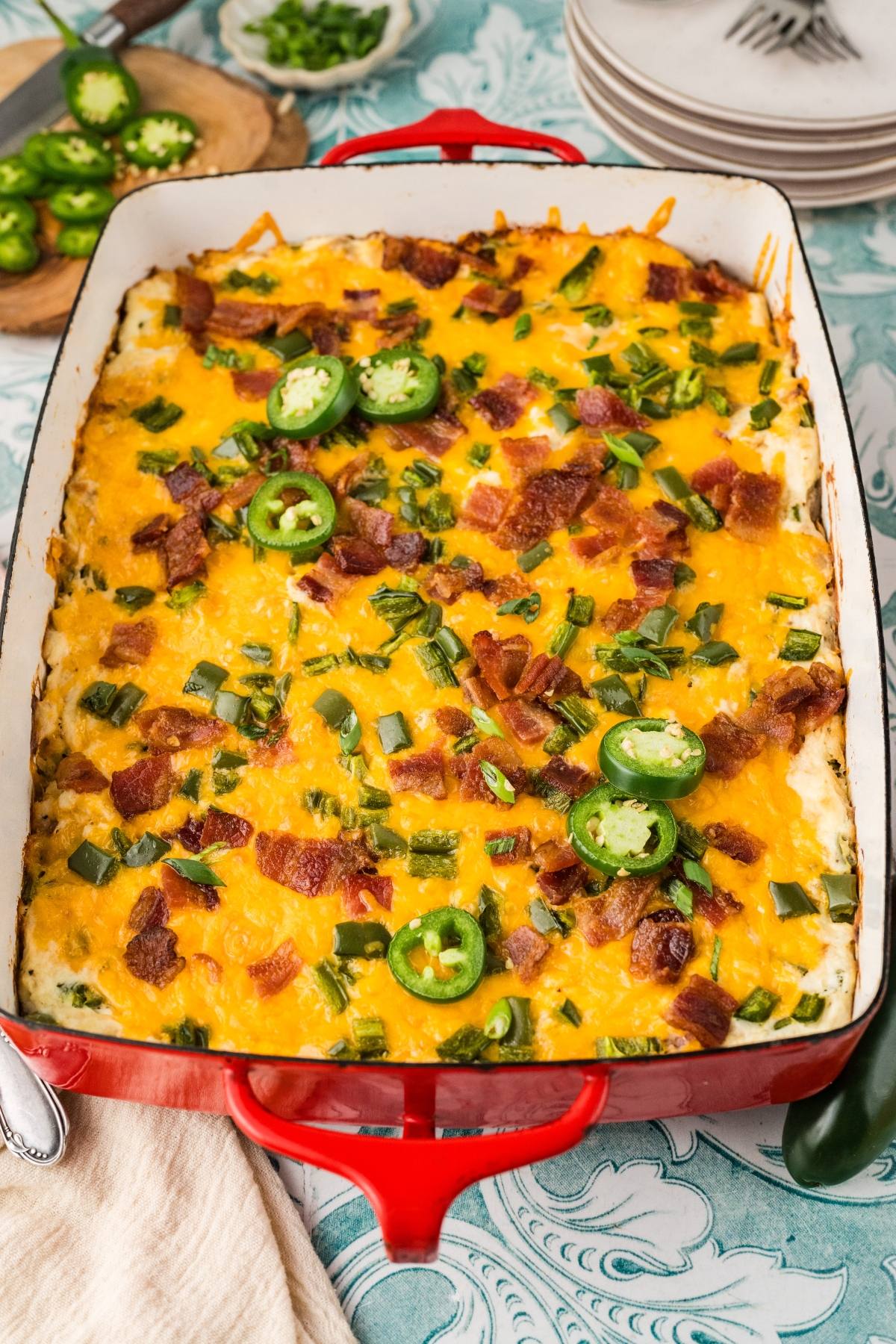 Baked cheesy casserole topped with bacon, green onions, and sliced jalapeños in a red baking dish.