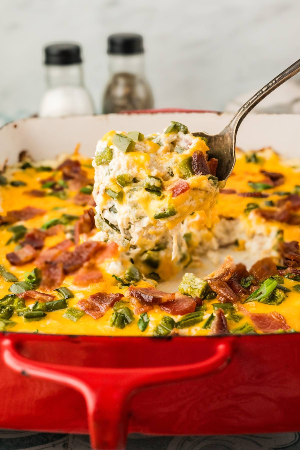 A spoon lifts cheesy, bacon-topped casserole from a red baking dish; salt and pepper shakers in background.