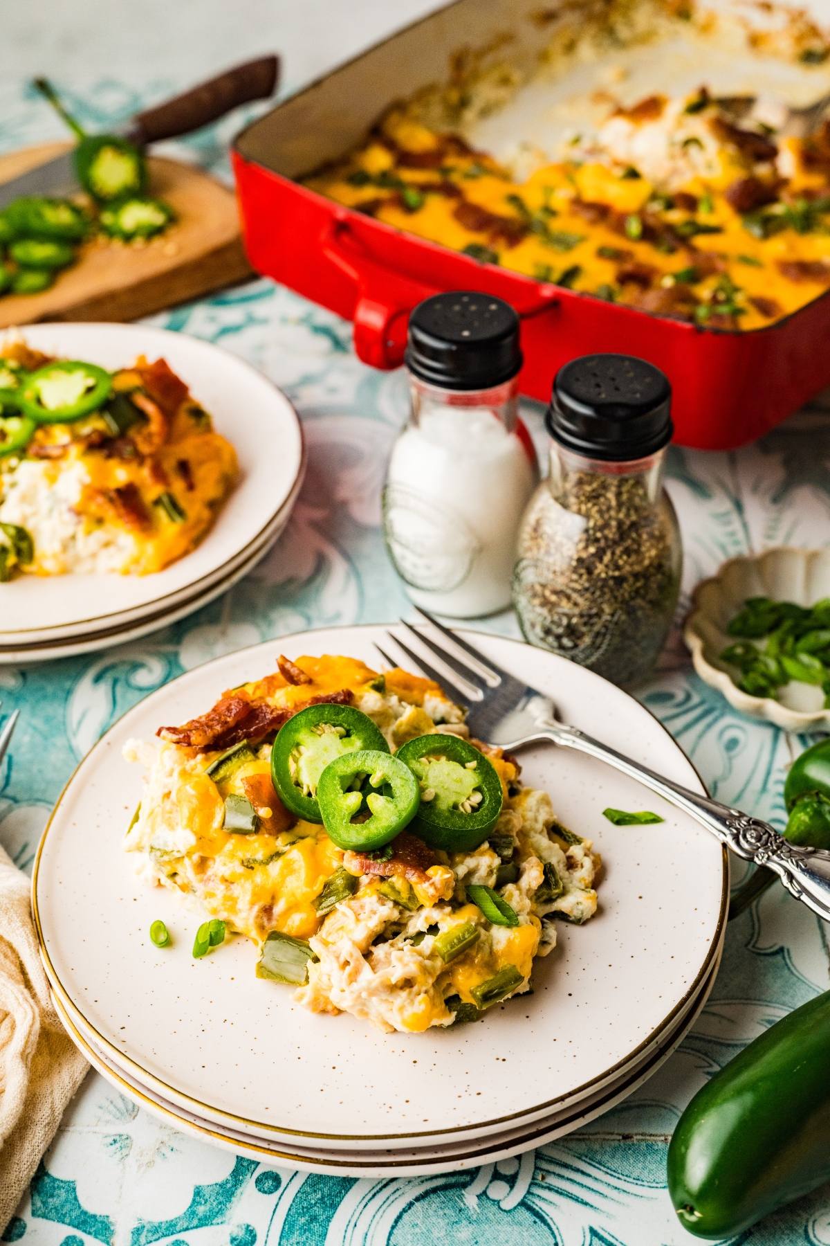 A plate of cheesy casserole topped with jalapeño slices, with a fork and a baking dish in the background.