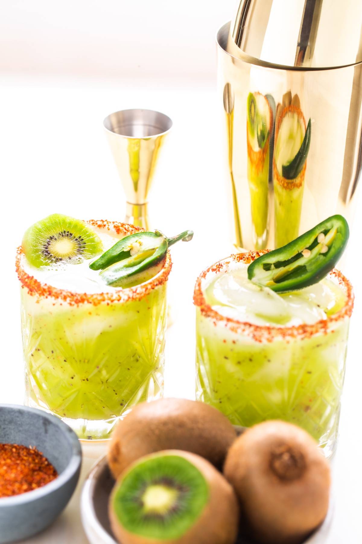 Two kiwi cocktails with chili-rimmed glasses, garnished with kiwi and jalapeño slices, beside fresh kiwis—this Kiwi Jalapeño Margarita is the best spicy margarita for bold flavor lovers.