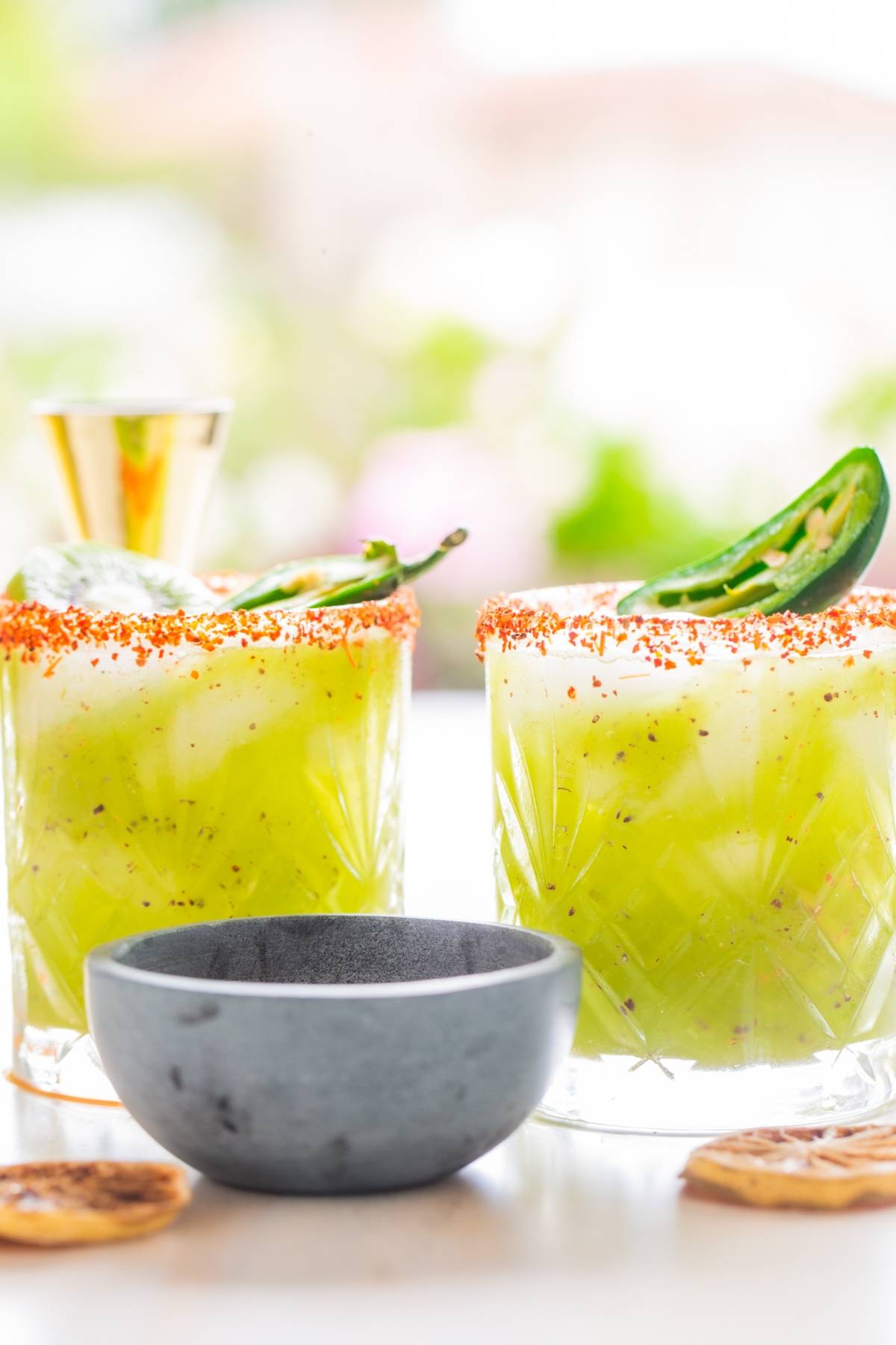 Two green cocktails made with kiwis, featuring chili powder rims and jalapeño slices, sit beside a small gray bowl and lime slices—a zesty margarita recipe for those craving the best spicy margarita experience.