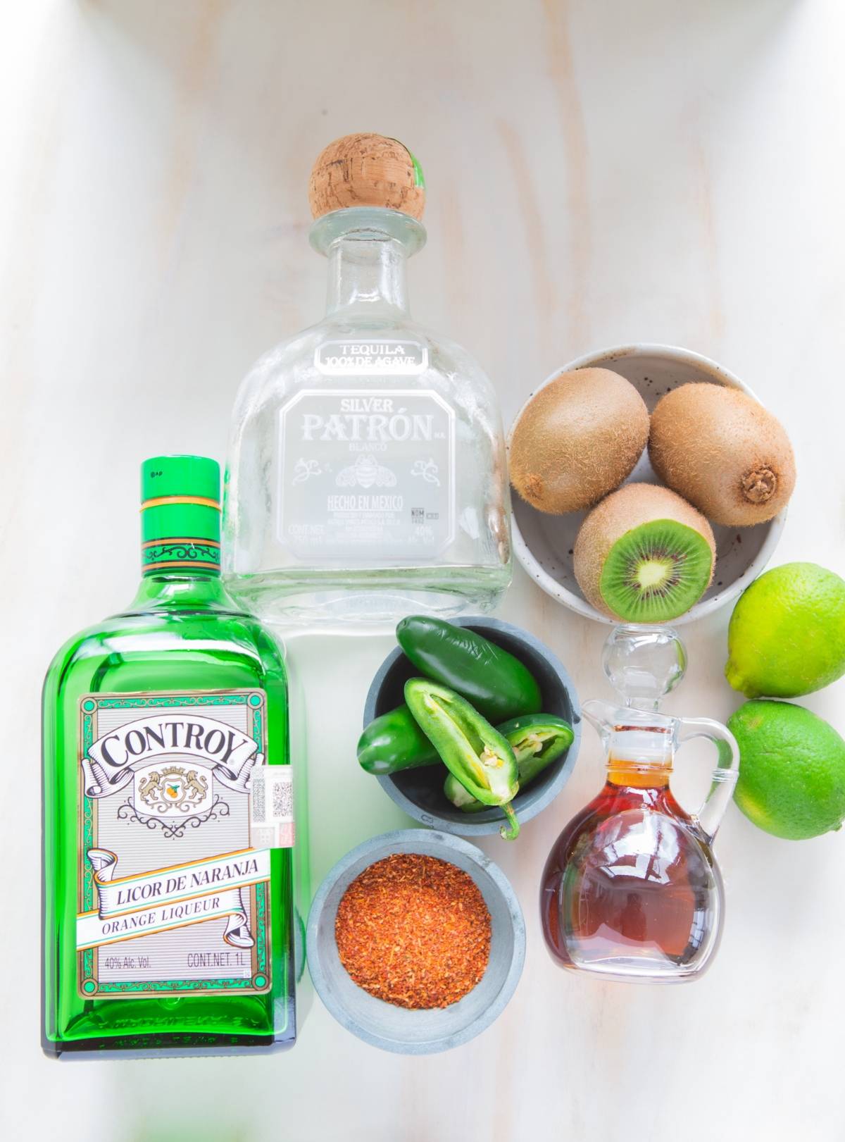 Bottles of tequila and orange liqueur, jalapeños, kiwi, limes, chili powder, and syrup on a white surface—perfect ingredients for a zesty margarita recipe like the Kiwi Jalapeño Margarita.