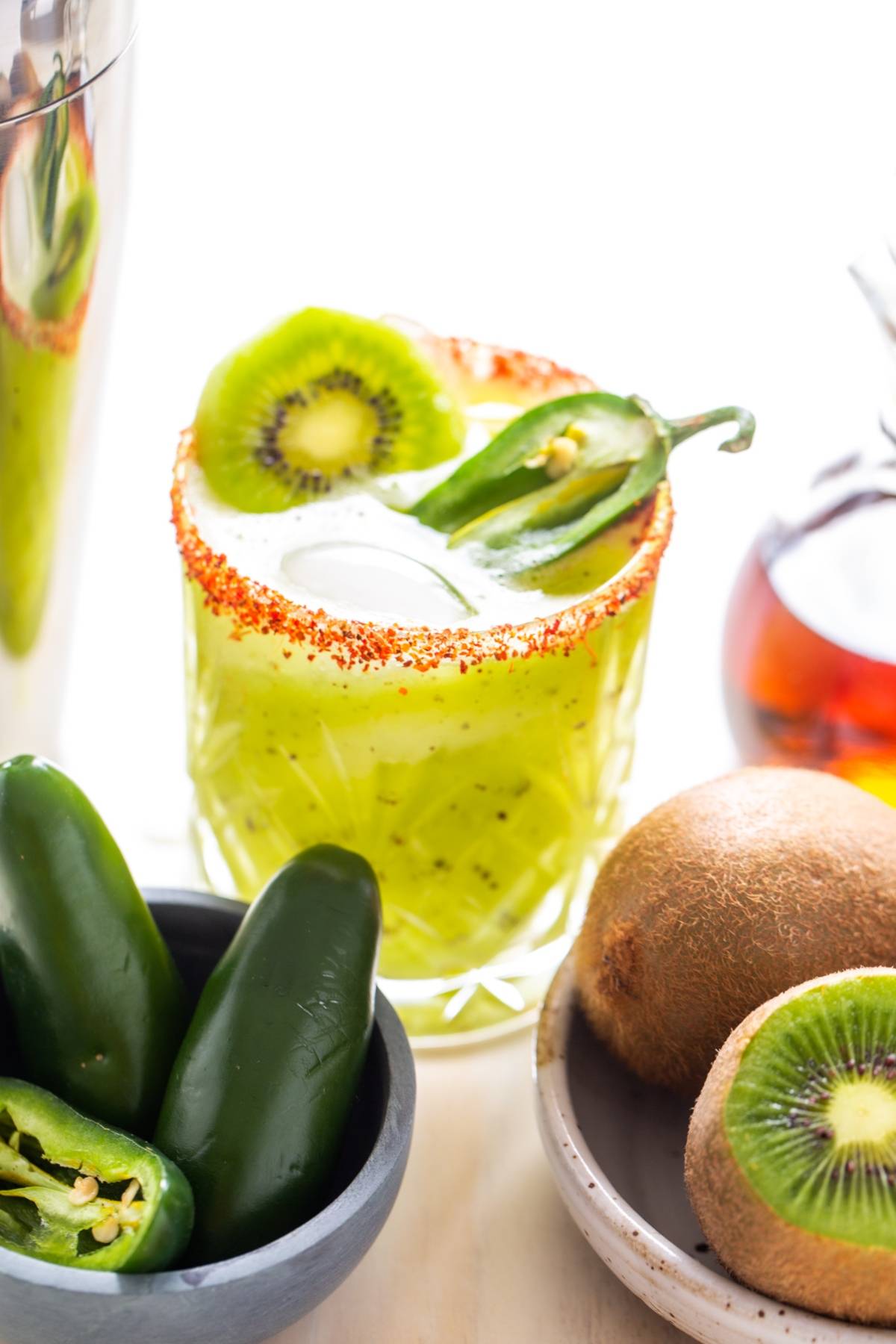 A zesty margarita recipe featuring a glass of kiwi cocktail with chili rim, garnished with kiwi and jalapeño, sits beside fresh kiwis and jalapeños.