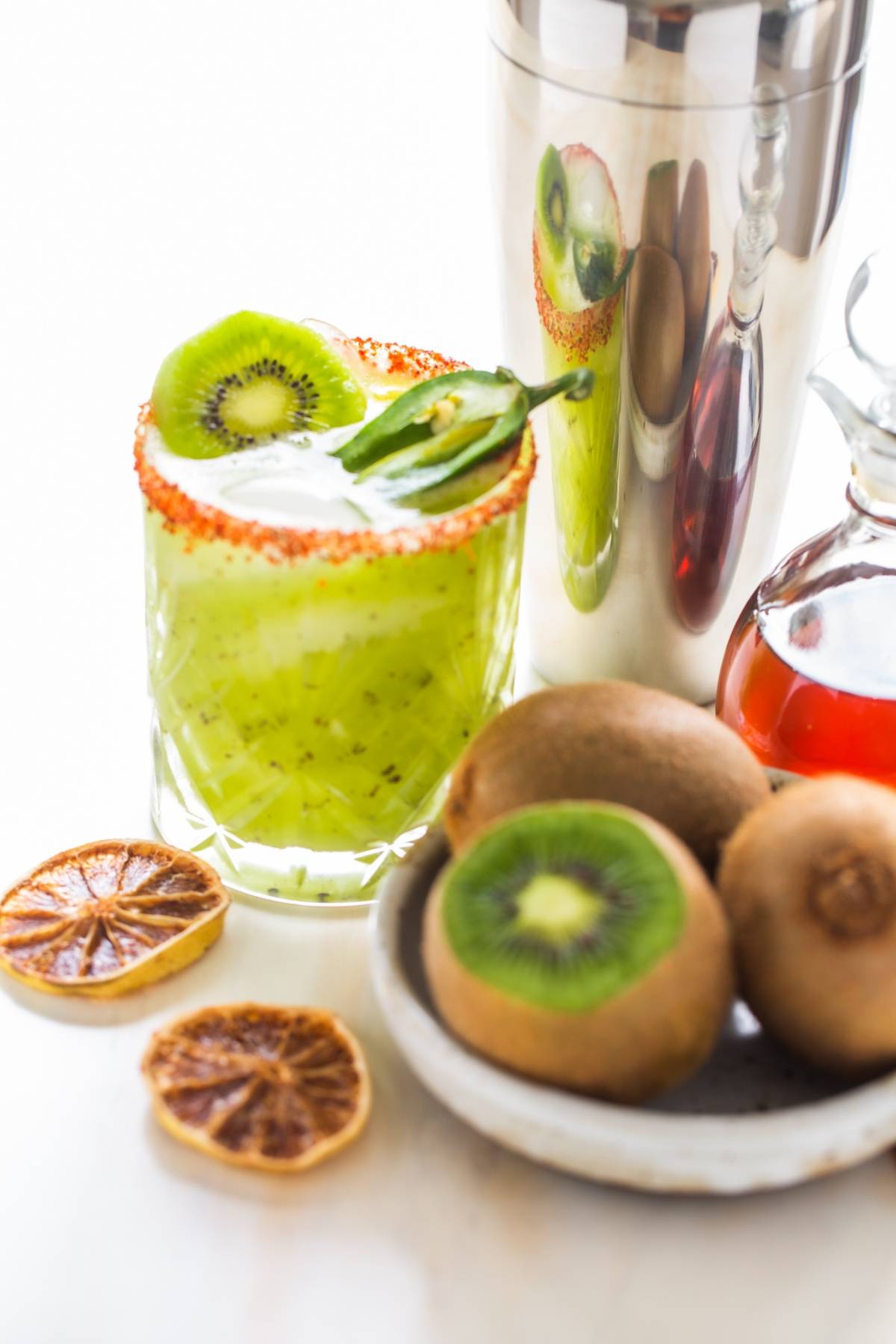 Green Kiwi Jalapeño Margarita in a glass with chili rim, fresh kiwis, dried citrus, syrup, and a cocktail shaker nearby—the best spicy margarita made with tequila and orange liqueur.