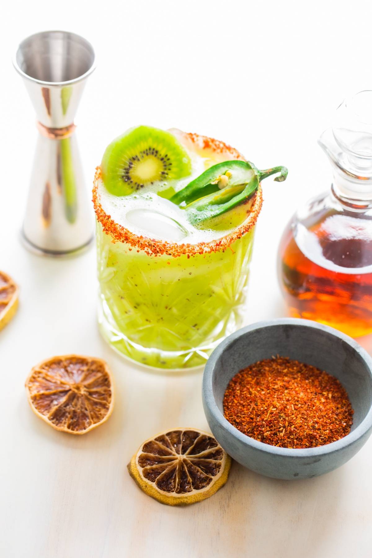 A Kiwi Jalapeño Margarita with a chili rim sits beside dried citrus, chili powder, and a jigger—a vibrant cocktail made with kiwis that could be the best spicy margarita for adventurous palates.