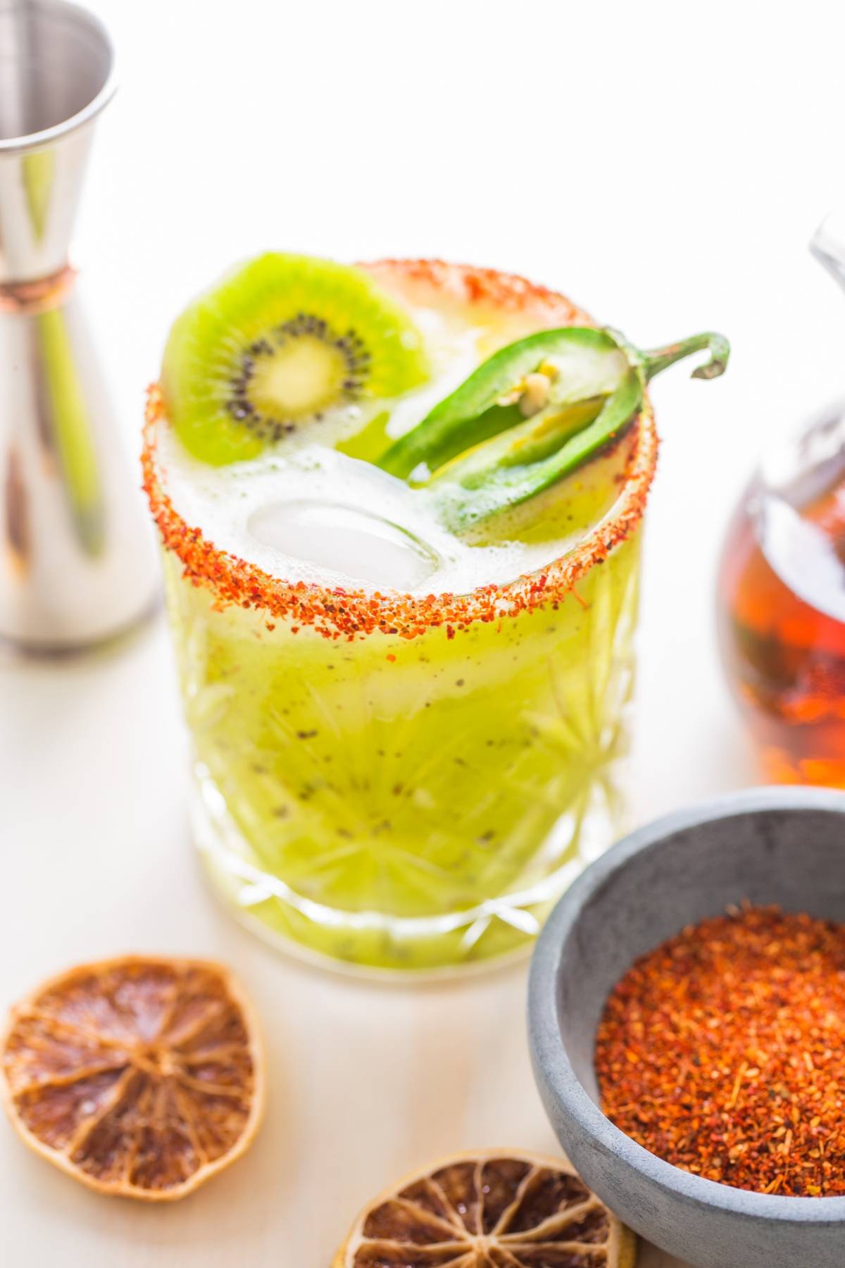 A green Kiwi Jalapeño Margarita with ice, kiwi slice, jalapeño, and chili powder rim, surrounded by dried citrus and spices—the best spicy margarita for those who crave a zesty twist.