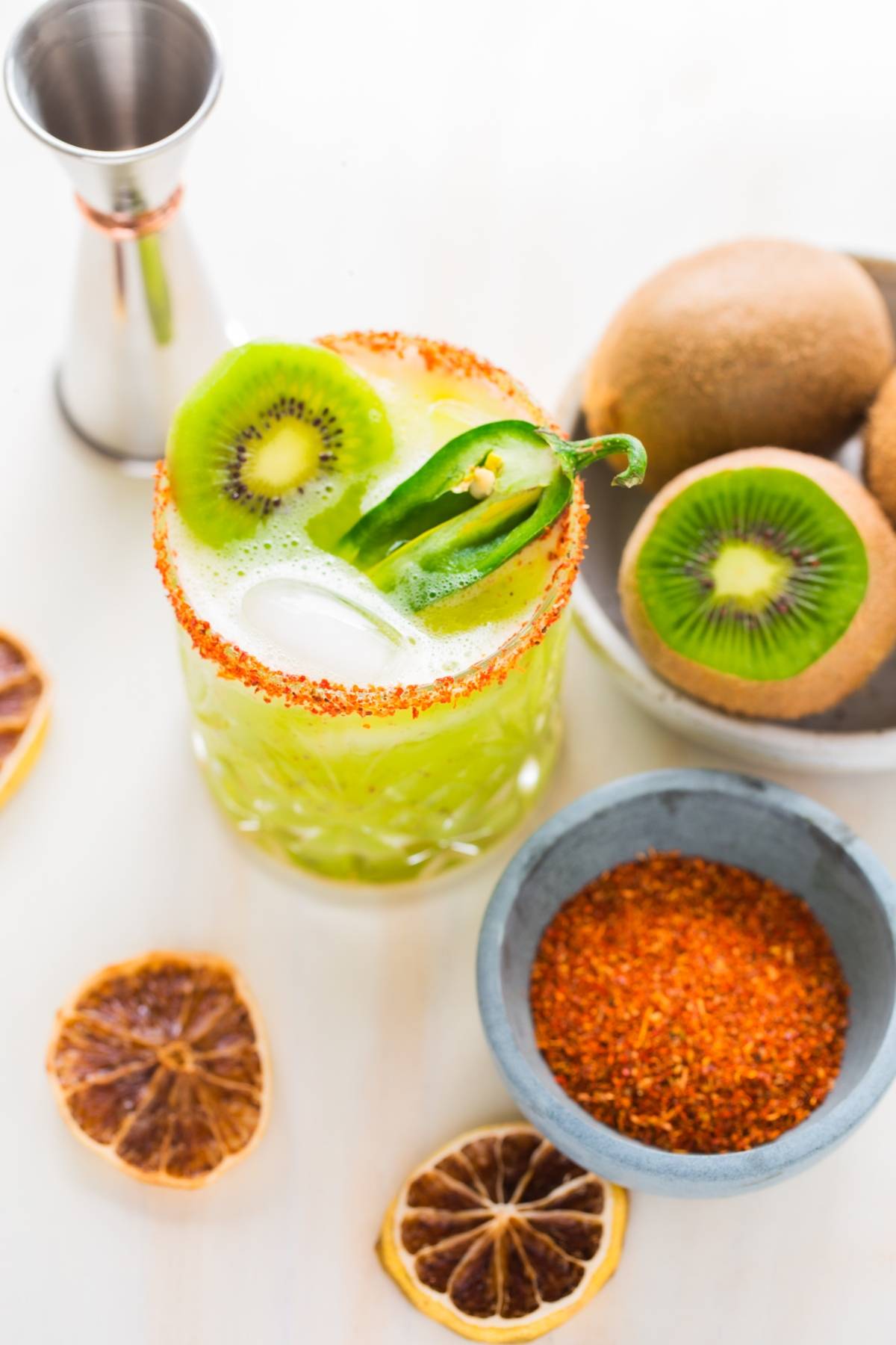 A green Kiwi Jalapeño Margarita with ice, chili rim, and jalapeño garnish—this cocktail using tequila and orange liqueur is surrounded by kiwi, dried lemon, and spices. Enjoy the best spicy margarita experience!.