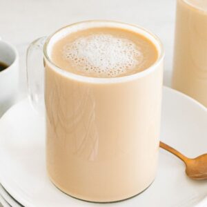 A glass mug of creamy latte breve sits on a white plate with a spoon, next to a cup of black coffee.