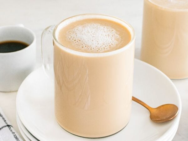 A glass mug of creamy latte breve sits on a white plate with a spoon, next to a cup of black coffee.