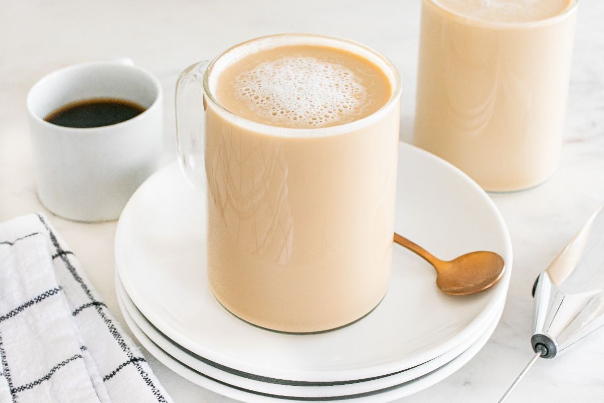 A mug of frothy latte breve on stacked plates with a spoon, next to a cup of black coffee and a napkin.