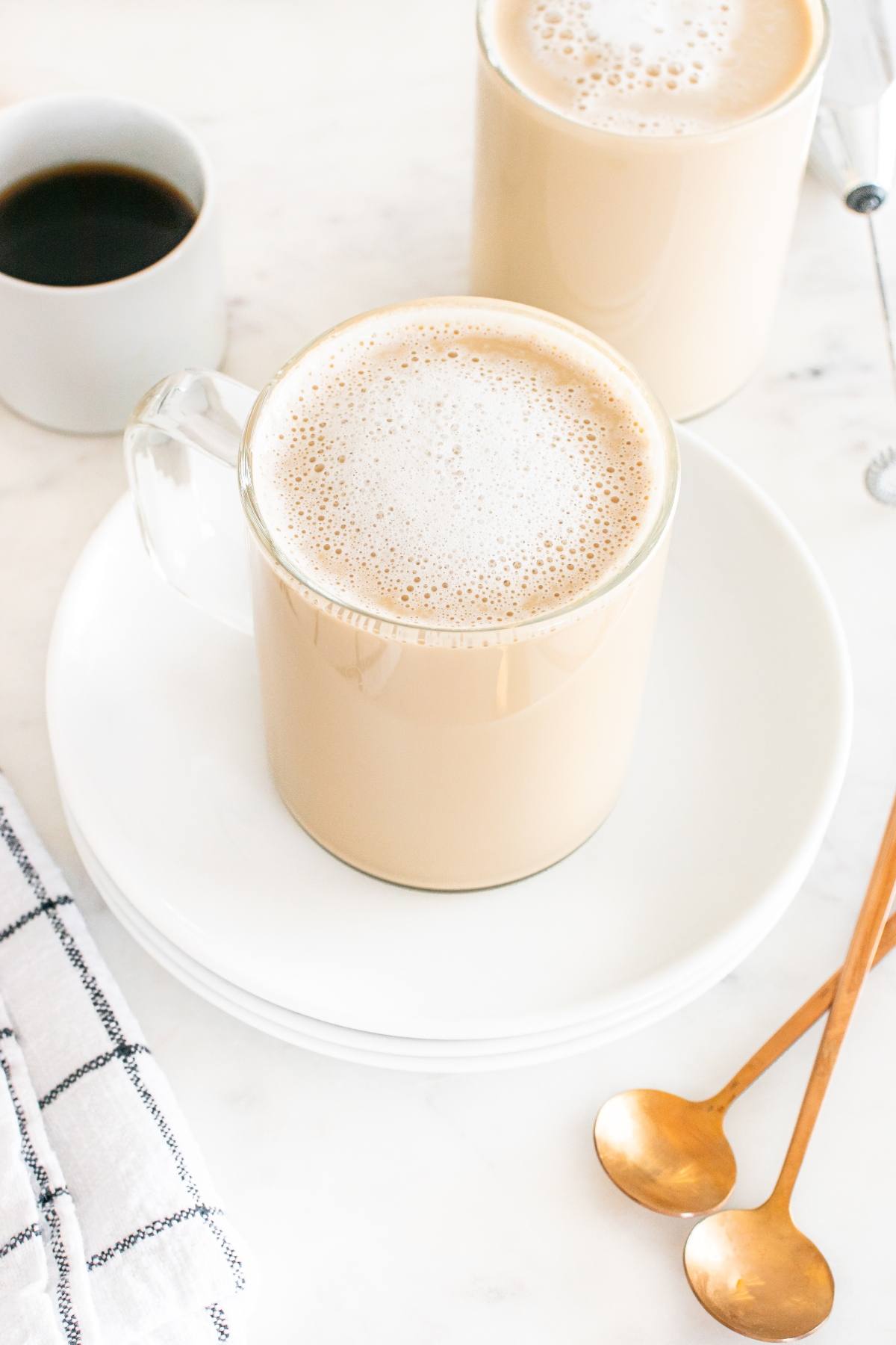 Clear glass mug of frothy coffee on a white plate, with gold spoons and a cup of black coffee nearby.