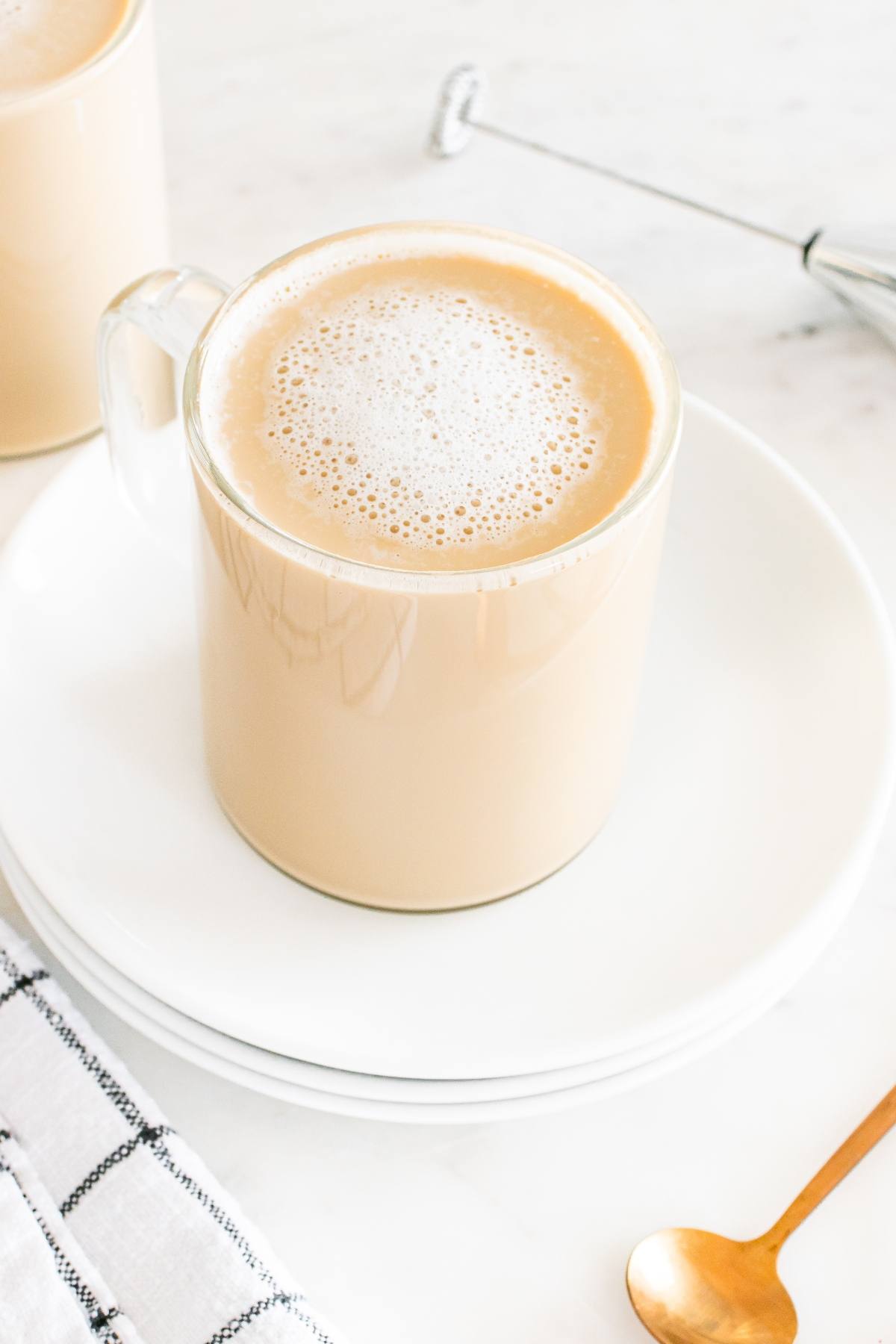 A glass mug of frothy coffee sits on white plates with a gold spoon and a plaid napkin nearby.