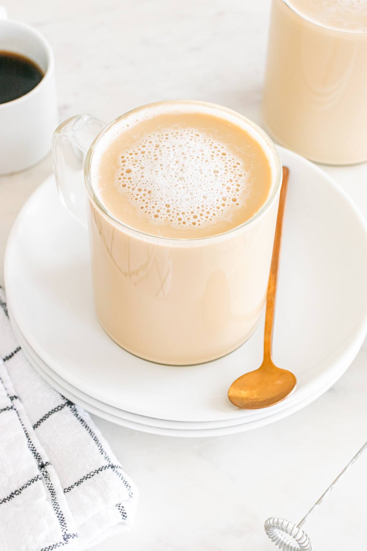 A glass mug of frothy coffee on a white plate with a gold spoon, next to a black-and-white napkin.