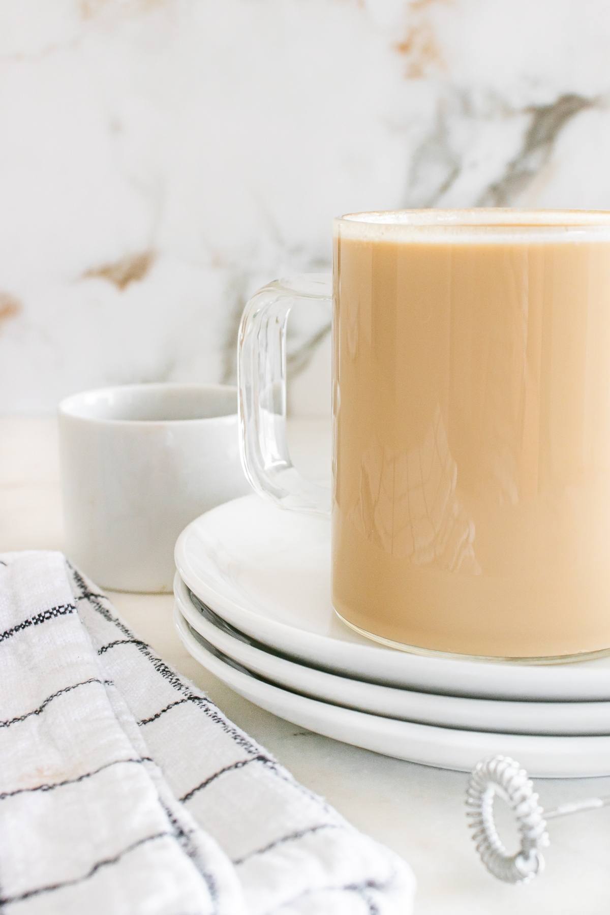 A glass mug of coffee with milk on stacked plates beside a small white cup and checkered cloth.