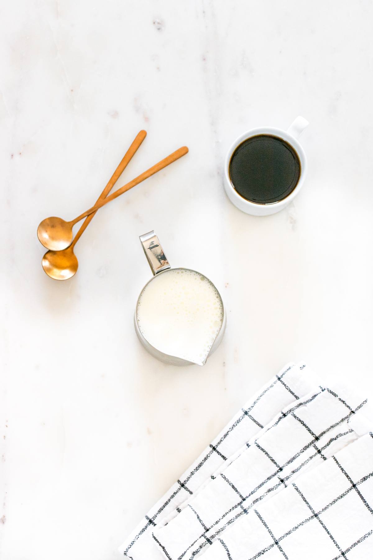 Cup of black coffee, a jug of milk, two golden spoons, and a checkered cloth on a white surface.